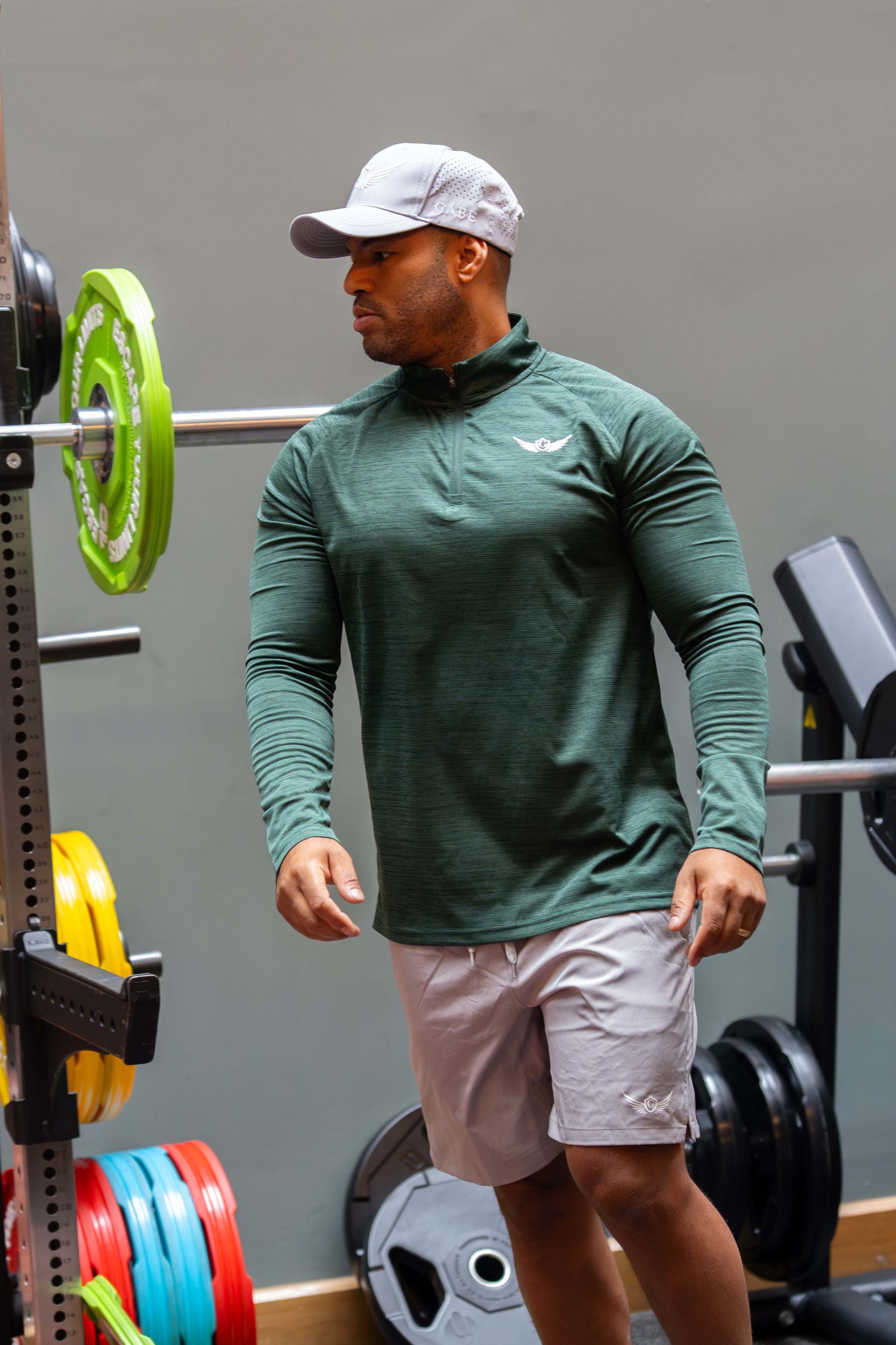 Man in green athletic shirt and grey shorts standing next to a barbell in a gym.
