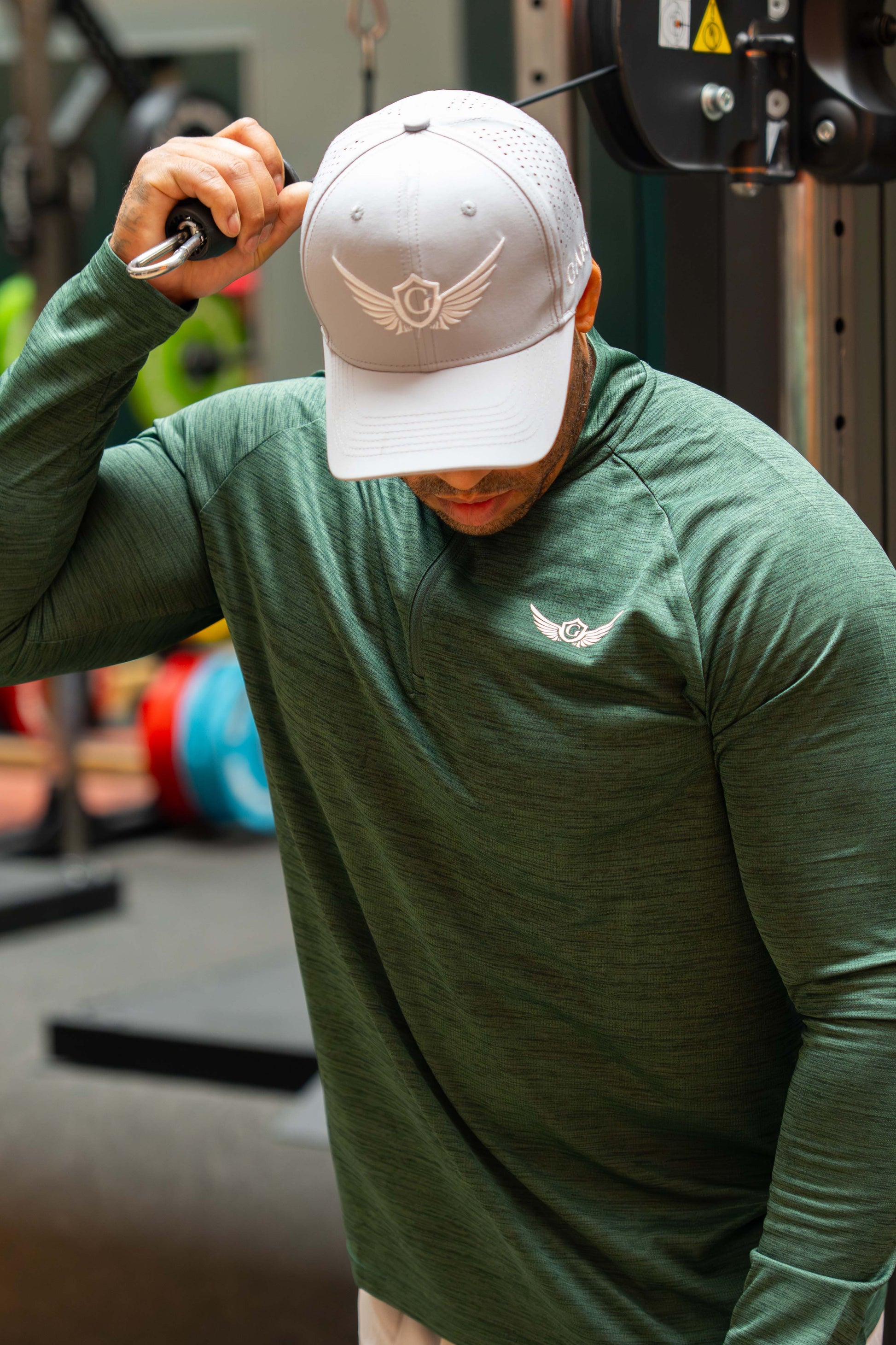 Man in a gym wearing a green long-sleeve shirt and a grey cap with a logo.
