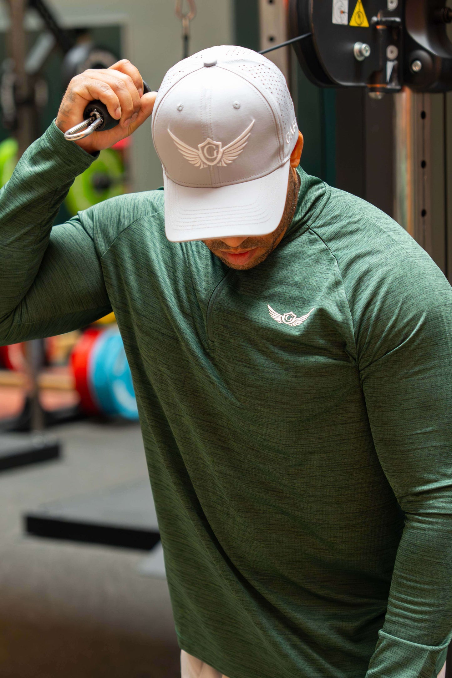 Man in a gym wearing a green long-sleeve shirt and a grey cap with a logo.