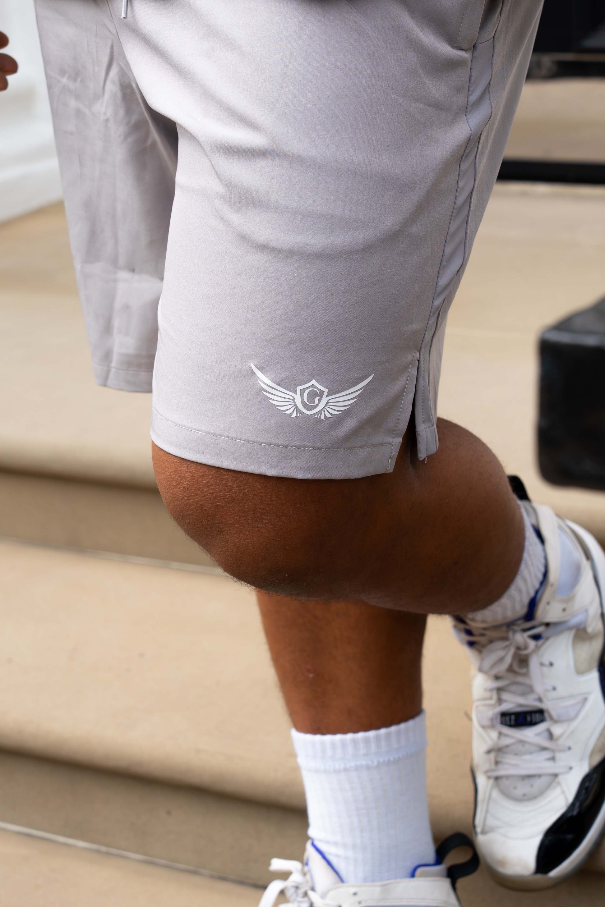 Person wearing grey shorts with a logo, white socks, and sneakers on a neutral background