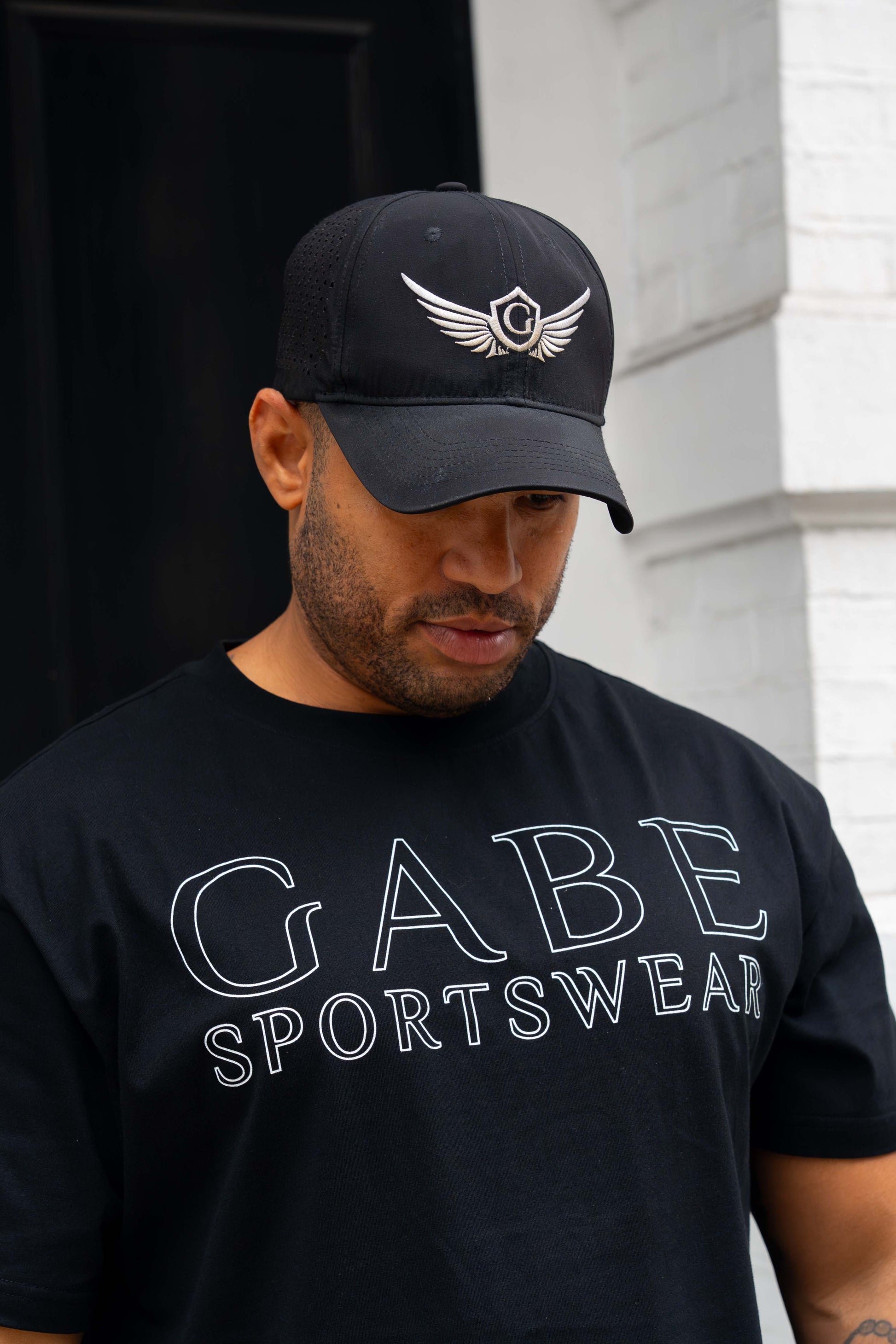 Man wearing a black cap and black 'GABE SPORTSWEAR' t-shirt against a white wall.