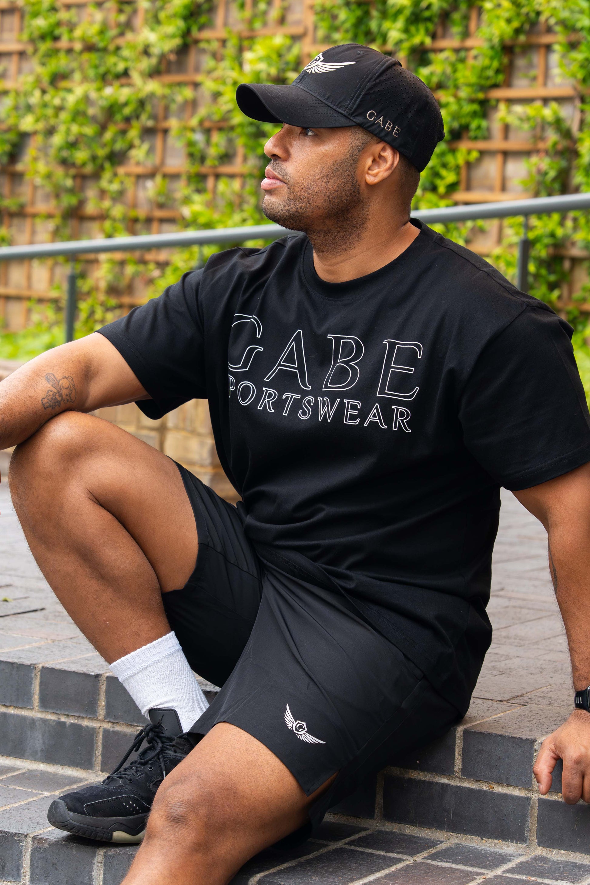 Man wearing a black 'GABE Sportswear' t-shirt and shorts sitting outdoors.