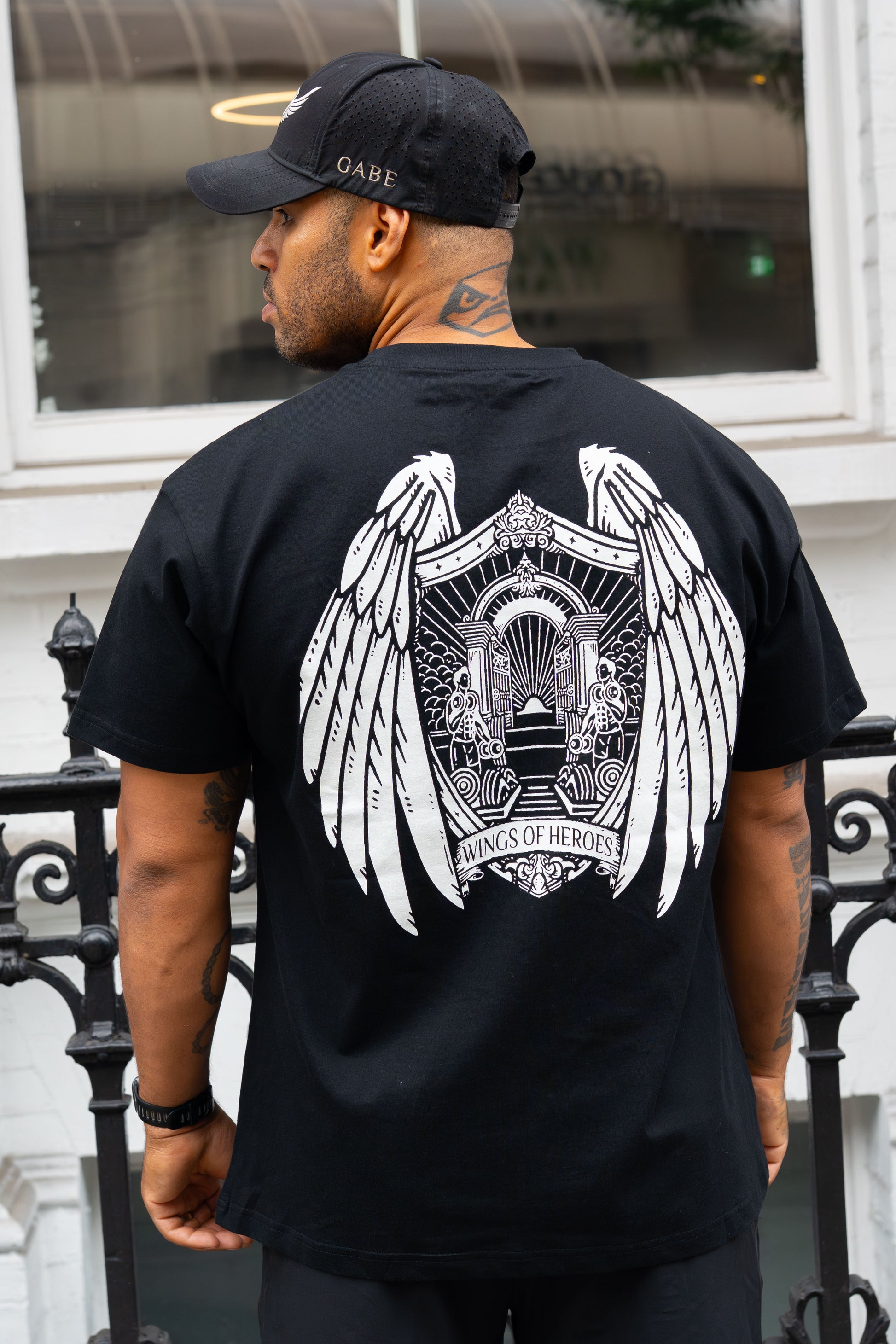 Man wearing a black t-shirt with a white graphic design on the back, standing in front of a building.