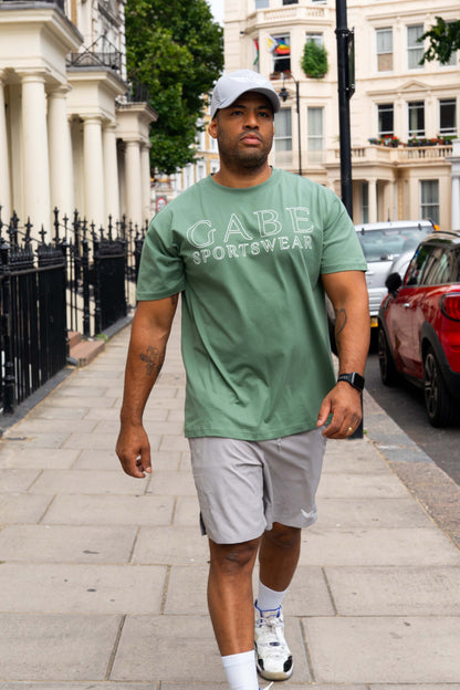 Man walking on a sidewalk wearing a green t-shirt with 'GABE SPORTSWEAR' text.